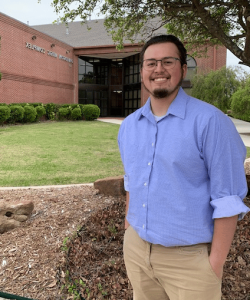 A smiling man in a blue shirt standing in front of a brick building at Howard Payne University, with a sign that reads "education service complex. | HPU